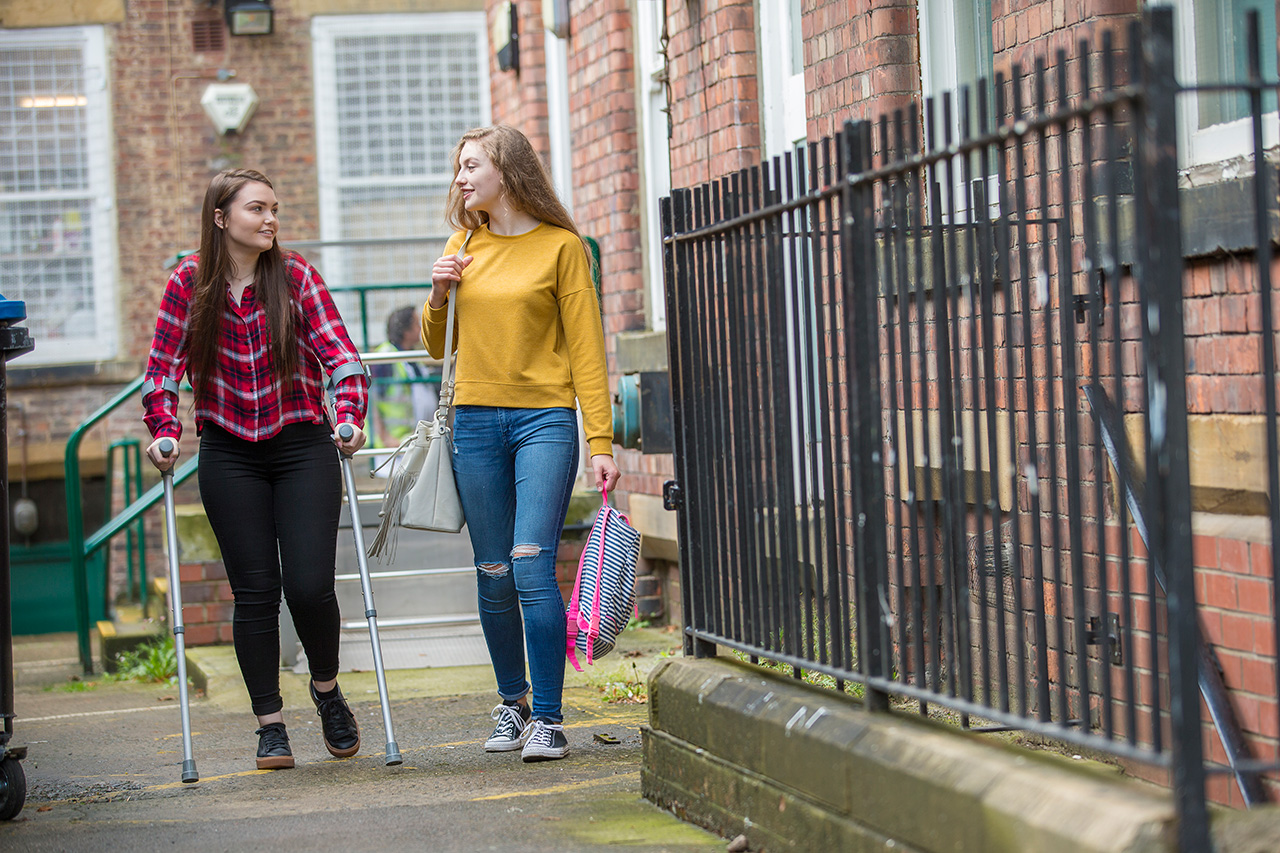 Two young women walking past a brick building, the woman wearing a flannel shirt is walking with crutches