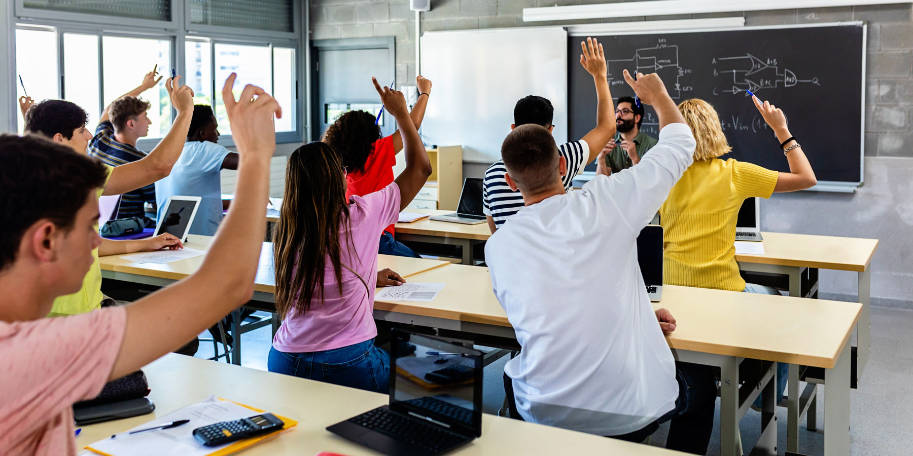 Diverse group of high school students hands raised in classroom setting.