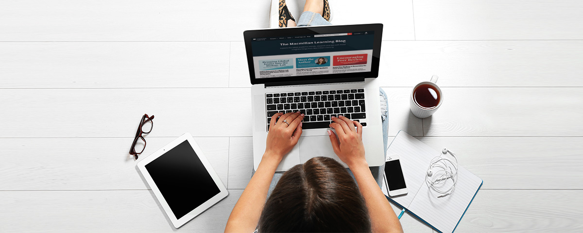 A photograph of a top view of a girl who is seated on the floor and working on the laptop placed on her lap. A tablet, a pair of spectacles, a coffee mug, a phone, a notebook, and earplugs are scattered around her.