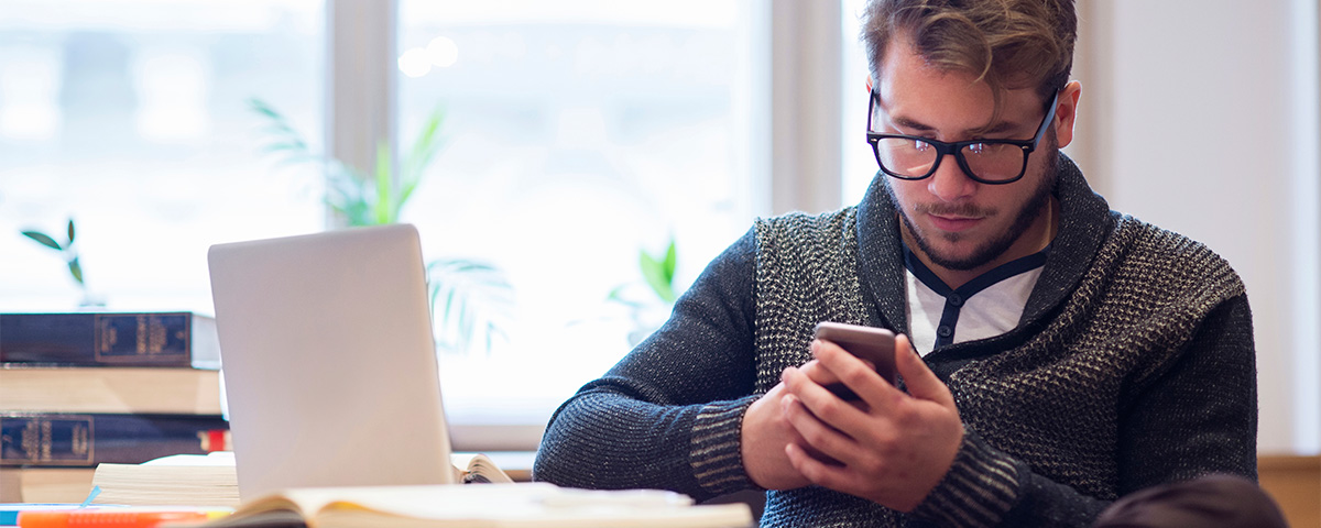 Photograph of man looking at phone next to laptop and pile of books.