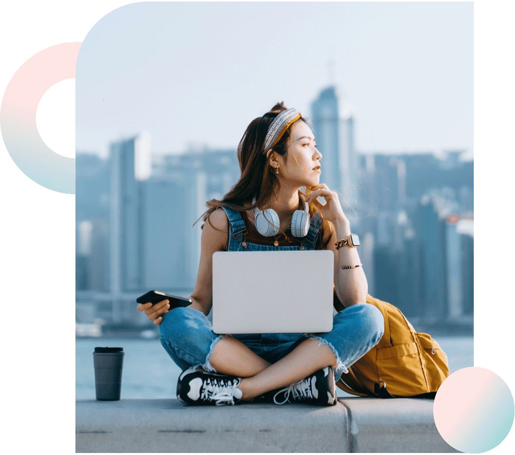 Beautiful young Asian woman sitting cross-legged by the promenade, against urban city skyline. She is wearing headphones around neck, using smartphone and working on laptop, with a coffee cup by her side. Looking away in thought.