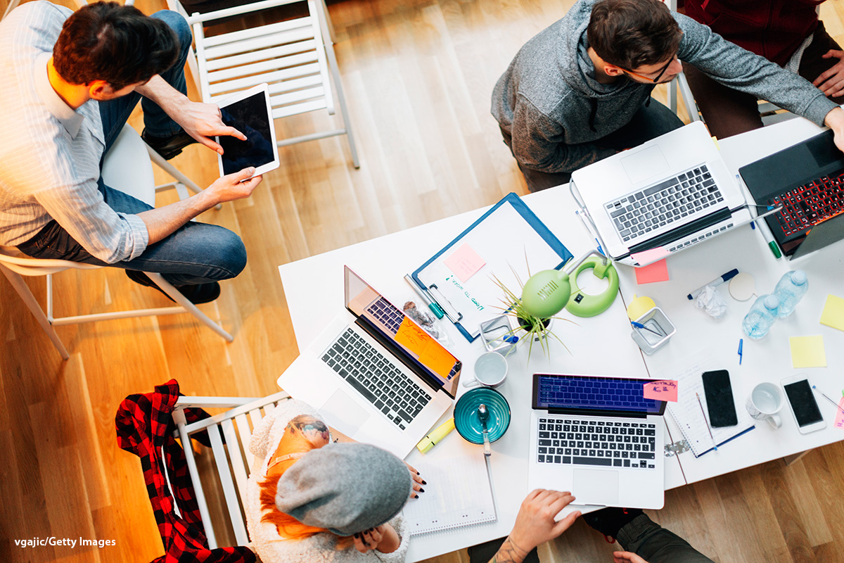 An aerial view photograph shows four individuals working. Three of them are seated by a desk and using multiple laptops, while the fourth is sitting on a chair and using a tablet. 