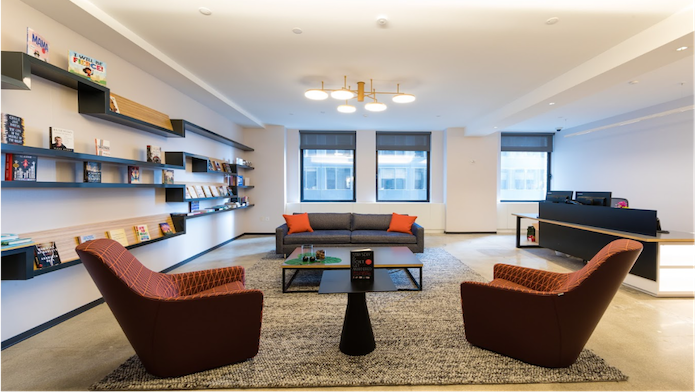 A photograph shows a well-lit office room set up with two lounge chairs placed on the floor. A center table is placed in between. A sofa, a few books on book racks, and a conference table are shown in the background. Three windows are shown in that room.