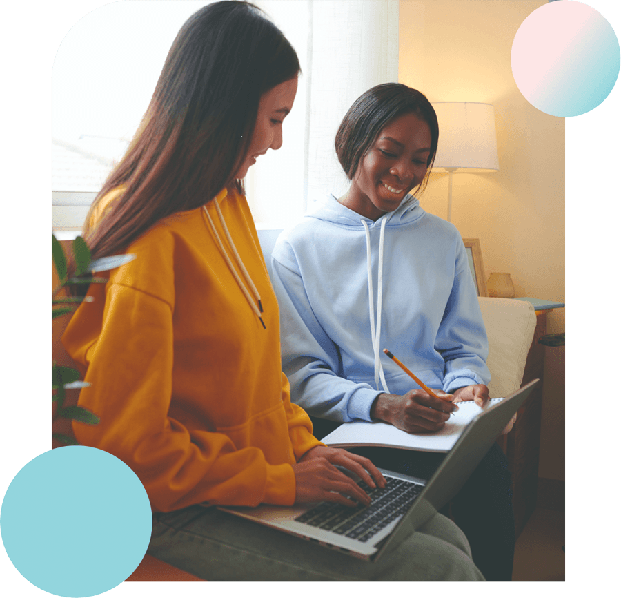 A photograph shows two women of different ethnicities seated on a couch and chatting casually. One woman is seen holding a laptop, while the other has a pen and notebook and taking down notes.