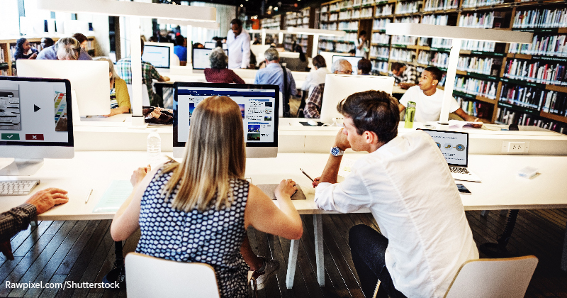 A photograph shows a college library set up with books arranged on the book racks. Several individuals are sitting with desktop computers and laptops at the tables.