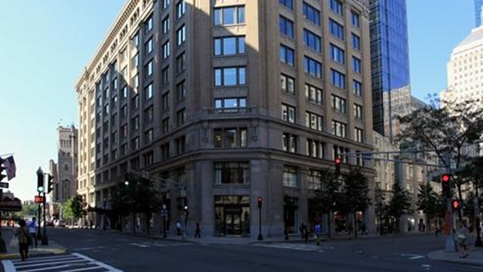 A photograph shows a huge office building with glass doors and windows at the side of a wide road with traffic lights. People are seen walking on the road. A few trees are shown on the roadside. 