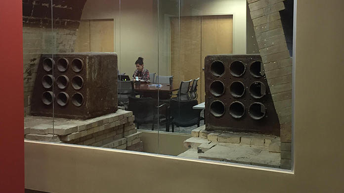 A photograph shows a well-lit office set-up. A girl is sitting at a conference table in a room with glass walls. The interior shows two large square blocks each with nine holes and placed on the stacked stone bricks.